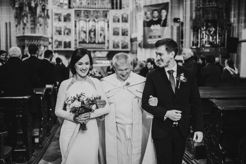 ciernobiela svadobna fotografia svadobneho paru vychadzajuceho z kostola s fararom